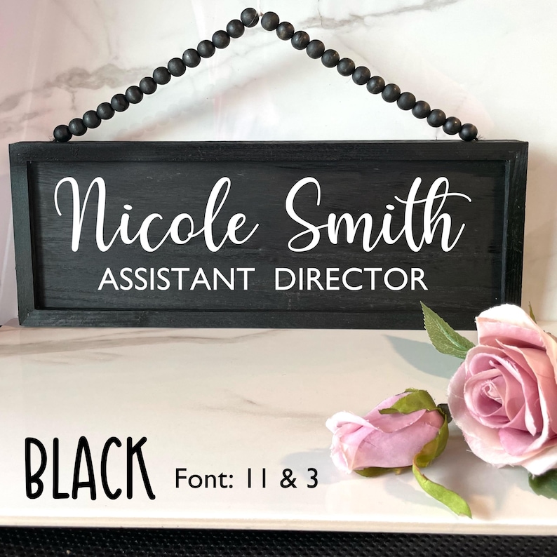 Peut inclure: Panneau en bois noir avec le nom "Nicole Smith" et le titre "ASSISTANT DIRECTOR" en lettres blanches. Le panneau est suspendu &agrave; une cha&icirc;ne de perles noires. Le mot "BLACK" et les d&eacute;tails de la police sont en bas. Des roses roses sont au premier plan.