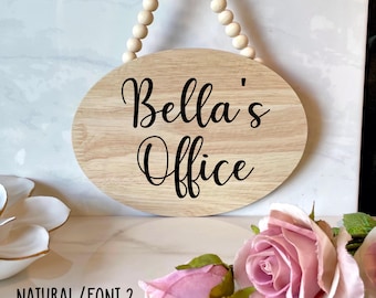 May include: A natural wood oval sign with the text "Bella's Office" in black cursive font. The sign is hanging from a beaded string.