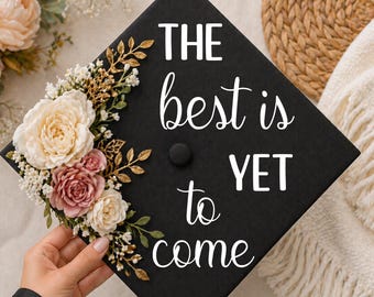 May include: Black graduation cap decorated with white script that reads "THE best is YET to come." The cap is adorned with a floral arrangement of cream, pink, and burgundy roses, with gold and green accents. The font is 5/14.