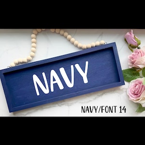 May include: A navy blue wooden sign with the word "NAVY" in white, bold letters. The sign is rectangular and has a dark blue frame. A string of wooden beads and pink roses are in the background. The text "NAVY/FONT 14" is at the bottom.