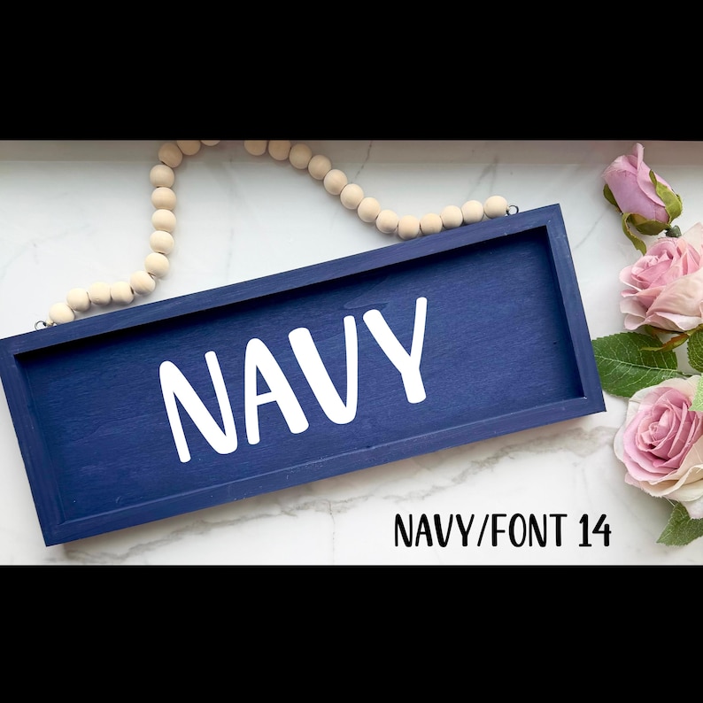 May include: A navy blue wooden sign with the word "NAVY" in white, bold letters. The sign is rectangular and has a dark blue frame. A string of wooden beads and pink roses are in the background. The text "NAVY/FONT 14" is at the bottom.