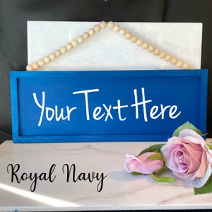 Peut inclure: Une enseigne en bois bleu marine avec le texte "Your Text Here" en &eacute;criture blanche. L'enseigne est encadr&eacute;e et comporte une guirlande de perles en bois en haut pour l'accrocher. Les mots "Royal Navy" sont &eacute;crits en bas.