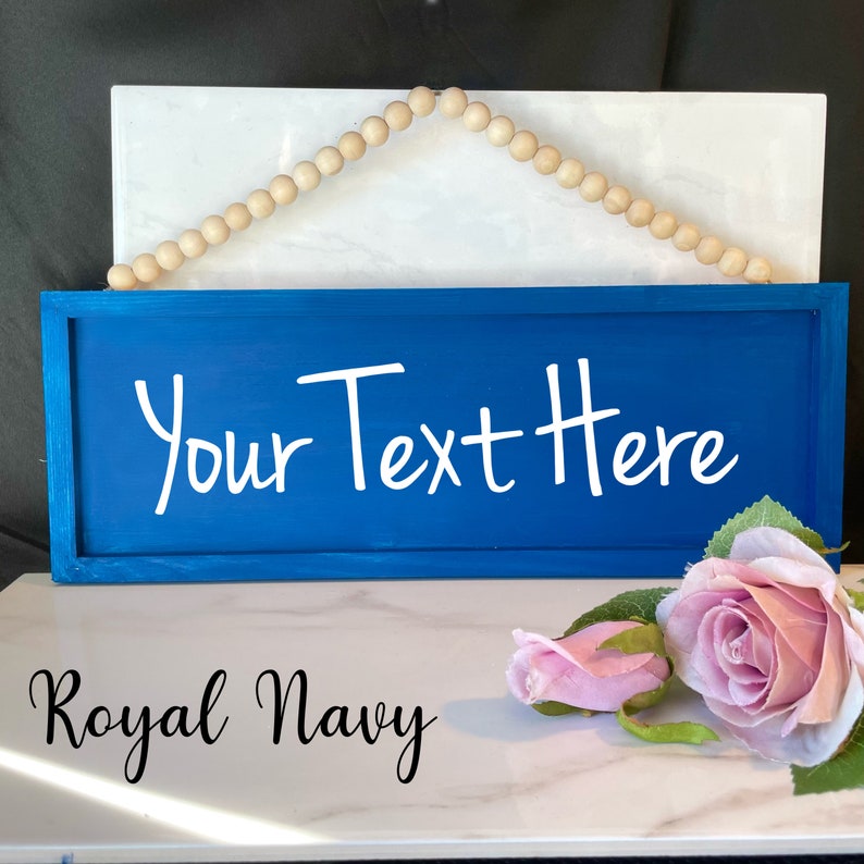 Peut inclure: Une enseigne en bois bleu marine avec le texte "Your Text Here" en &eacute;criture blanche. L'enseigne est encadr&eacute;e et comporte une guirlande de perles en bois en haut pour l'accrocher. Les mots "Royal Navy" sont &eacute;crits en bas.