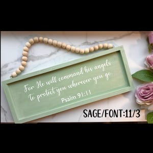 May include: Sage green wooden sign with white lettering that reads "For He will command his angels to protect you wherever you go. Psalm 91:11." The sign is rectangular with a beaded wooden hanger. The text "SAGE/FONT:11/3" is in the bottom right corner.