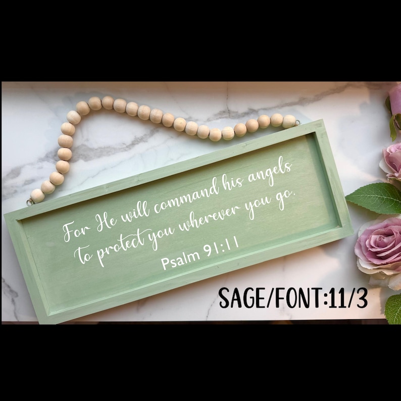 May include: Sage green wooden sign with white lettering that reads "For He will command his angels to protect you wherever you go. Psalm 91:11." The sign is rectangular with a beaded wooden hanger. The text "SAGE/FONT:11/3" is in the bottom right corner.