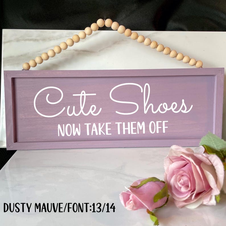 May include: A rectangular, dusty mauve wooden sign with the words "Cute Shoes NOW TAKE THEM OFF" in white script. The sign is suspended by a string of wooden beads. Pink roses and buds are in the foreground.