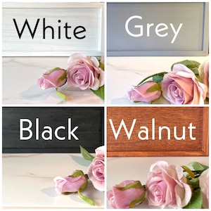 May include: Four wooden signs with different paint colors: white, grey, black, and walnut. Each sign has the color name written in white text. Pink roses are placed in front of each sign.