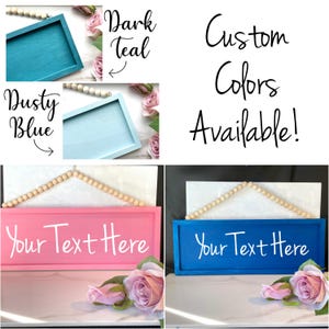 May include: A collection of wooden signs in various colors, including dark teal, dusty blue, pink, and blue. Each sign has a rectangular shape with the text "Your Text Here" in white. The signs are decorated with wooden beads and faux flowers. The text "Custom Colors Available!" is also present.