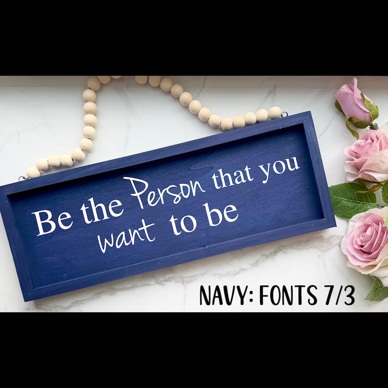 May include: A navy blue wooden sign with the text "Be the Person that you want to be" in white script. The sign is rectangular and framed. A string of wooden beads and pink roses are in the background.