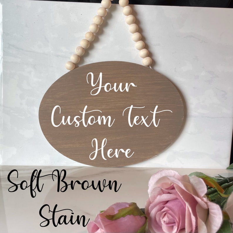 Pu&ograve; includere: Un cartello ovale in legno marrone con testo bianco che recita "Your Custom Text Here". Il cartello &egrave; appeso a una corda di perline di legno. Il cartello &egrave; su uno sfondo di marmo bianco con rose rosa in primo piano. Il testo "Soft Brown Stain" &egrave; scritto sotto il cartello.