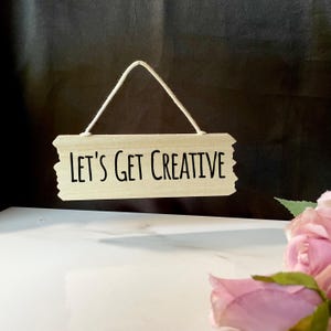 May include: A wooden sign with a white string hanging from the top. The sign has the text "Let's Get Creative" written in black.