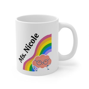 Könnte beinhalten: Weiße Keramik-Kaffeetasse mit einem Regenbogen und einem Cartoon-Gehirn mit Brille. Der Text "Ms. Nicole" steht auf der Tasse.