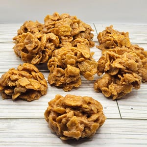 May include: Several clusters of golden-brown, crunchy treats, likely a type of no-bake cookie, are arranged on a light-colored wooden surface. The treats appear to be made with cereal and a sweet, sticky binder.