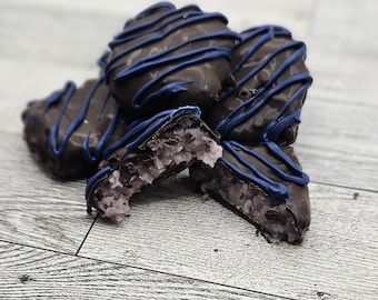 May include: A close-up of several chocolate-covered treats with a blue drizzle. One treat is broken open, revealing a light purple filling. The treats are arranged on a light-colored wooden surface.