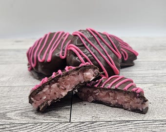 May include: Close-up of chocolate-covered treats with pink drizzle. One treat is cut in half, revealing a pink, coconut-based filling. Other treats are stacked in the background. The treats are on a light-colored wooden surface.