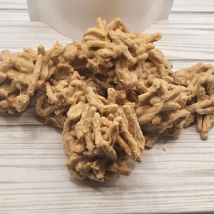 May include: A close-up of a pile of peanut butter patties, a type of candy, on a light wood surface. The patties are made with peanut butter, powdered sugar, and butter, and are shaped into small, irregular mounds.