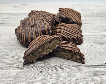 May include: A collection of chocolate-covered candies, some with a drizzle pattern. One candy is cut in half, revealing a light-colored, textured filling. The candies are arranged on a light-colored, wooden surface.