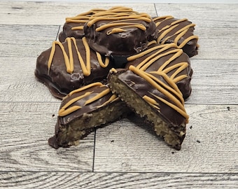 May include: A pile of chocolate-covered treats with peanut butter drizzle. One is cut in half, revealing a light-colored filling. The treats are arranged on a light-colored wooden surface. The chocolate coating is dark brown, and the drizzle is a light tan color.