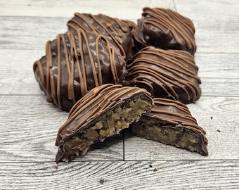 May include: Several chocolate-covered confections with a nutty interior are arranged on a light wood surface. The candies are drizzled with a darker chocolate, and one is cut in half, revealing the filling.