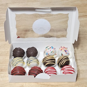 May include: A white box with a clear lid containing 10 chocolate-covered cake pops. The cake pops are decorated with different colors of sprinkles, stripes, and swirls.