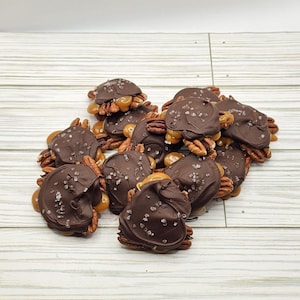 May include: A close-up of a pile of chocolate-covered caramel pecan clusters. The clusters are topped with sea salt and are arranged on a white wooden surface.