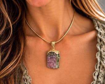 May include: A gold-toned snake chain necklace featuring a rectangular pendant with a raw, multi-colored gemstone. The stone displays shades of purple and green with a textured, crystalline surface, held by a gold-toned metal cap and bail.