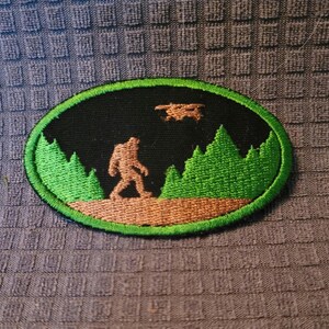 May include: Green and brown embroidered patch with a black background. The patch features a silhouette of a Bigfoot walking through a forest with a drone flying overhead.