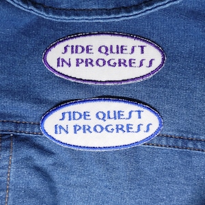 May include: Two oval-shaped embroidered patches attached to blue denim fabric. Both patches feature a white background with the text "SIDE QUEST IN PROGRESS" in a stylized font. The top patch has purple text and a purple border, while the bottom patch has blue text and a blue border.