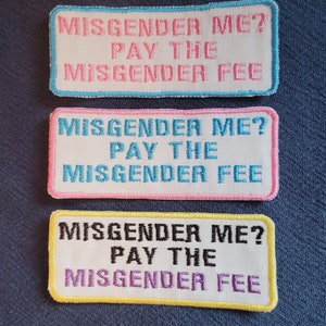 May include: Three embroidered patches with the text "Misgender Me? Pay the Misgender Fee" in white text on a white background. The patches have different colored borders: light blue, pink, and yellow.