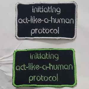 May include: Two rectangular embroidered patches with black fabric backgrounds. The top patch features white text reading "initiating act-like-a-human protocol" with a white border. The bottom patch features lime green text and a lime green border.