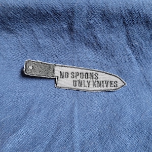May include: A gray and black knife-shaped patch with the text "NO SPOONS ONLY KNIVES" embroidered on it. The patch is set against a blue fabric background, creating a contrasting visual.