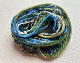 Puede incluir: Primer plano de un ovillo de hilo en tonos verdes, azules y blancos. El hilo está retorcido y enredado, creando un efecto colorido y texturizado.