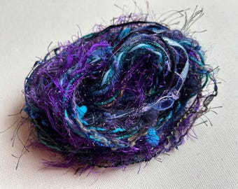 May include: A vibrant ball of yarn, predominantly purple with teal and black accents. The yarn has a textured, multi-strand appearance with metallic threads. Suitable for knitting, crochet, and other textile crafts.