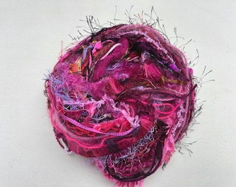 May include: A ball of colorful yarn in shades of pink, purple, and magenta. The yarn has a textured appearance with various fibers and fringe, creating a visually interesting and tactile craft supply.