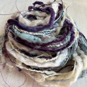 May include: A coiled bundle of decorative yarn featuring a mix of textures and colors including deep purple, cream, white, and soft blue. The yarn consists of various strands, some with a fuzzy, velvet-like appearance and others with thin metallic threads.