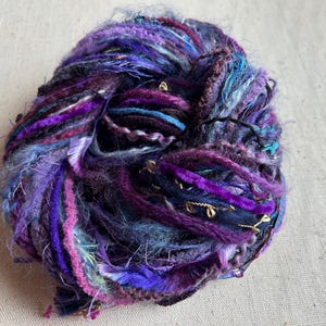 May include: A ball of art yarn featuring a mix of textures and fibers in shades of purple, blue, and magenta. The yarn includes fuzzy, velvet-like, and metallic gold strands twisted together to create a complex, multi-dimensional appearance.