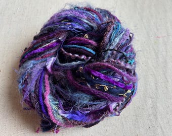 May include: A ball of art yarn featuring a mix of textures and fibres in shades of purple, blue, and magenta. The yarn includes fuzzy, velvet-like, and metallic gold strands twisted together to create a complex, multi-dimensional appearance.
