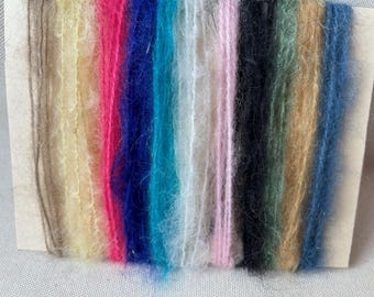 Puede incluir: Una colección de 12 hebras de hilo difuso de diferentes colores, mostradas en una tarjeta marrón. Los colores incluyen beige, amarillo, rosa, rojo, azul, azul verdoso, blanco, rosa claro, negro, verde, dorado y azul claro.