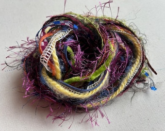 May include: A bundle of colourful yarn with a variety of textures, including fuzzy, smooth, and sparkly threads. The yarn is in shades of purple, yellow, green, blue, and orange.