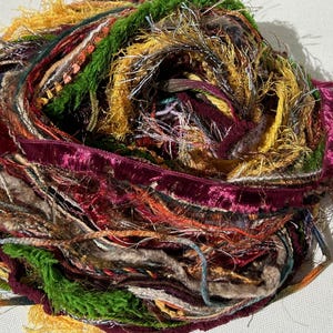 May include: A colorful ball of mixed yarn, featuring a variety of textures and hues. The yarn includes shades of green, yellow, burgundy, and pink, with some strands appearing metallic. The ball is tightly wound, showcasing the diverse materials.