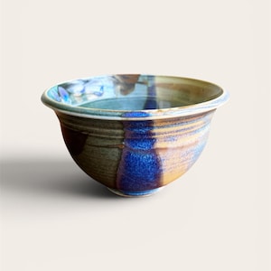 May include: A handmade ceramic bowl with a wide rim. The bowl features a blend of colors, including blue, brown, and yellow, with a glossy finish. The bowl's design is a mix of glazed and unglazed sections, creating a unique texture.