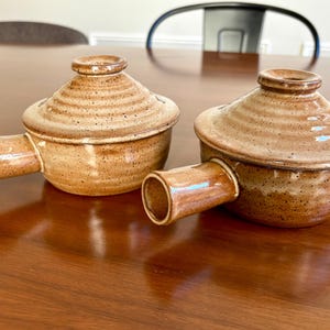 May include: Two brown ceramic lidded bowls with handles. The bowls have a speckled, textured finish and a round, slightly conical shape. The handles are cylindrical. The lids have a knob on top.