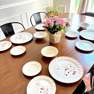 May include: A collection of cream-colored ceramic dinnerware featuring a swirling rim texture and a brown floral motif. The set includes various sizes of plates and bowls arranged on a wooden table, centered around a potted pink flower plant.