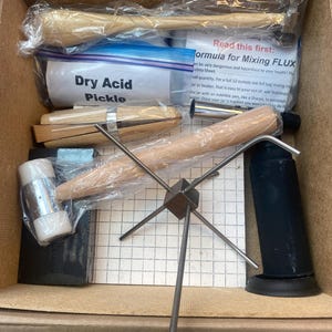 Può includere: Una scatola contenente vari strumenti per la creazione di gioielli. Include mazzuoli di legno, un contenitore con l'etichetta "Dry Acid Pickle", un supporto in metallo e altri oggetti avvolti. È visibile anche un foglio di carta con istruzioni.