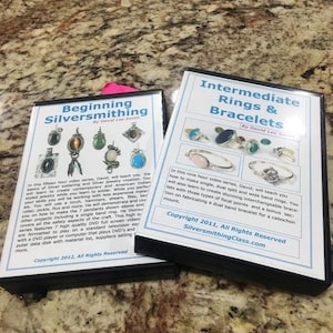 Pode incluir: Dois DVDs instrutivos sobre ourivesaria. Um é intitulado "Beginning Silversmithing" e o outro "Intermediate Rings & Bracelets". As capas apresentam imagens de joias e texto descrevendo o conteúdo e informações de direitos autorais.