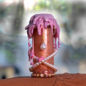 May include: A decorative, cylindrical object with a metallic rose gold finish, adorned with dripping pink wax. A pearl necklace and a gold chain with a pearl pendant wrap around the object. The base features a band with seashell accents.