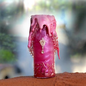 May include: A decorative candle with a pink and magenta color scheme. The candle has a dripping wax design at the top and is adorned with clear crystals and a jeweled pendant. The candle sits on a brown surface.
