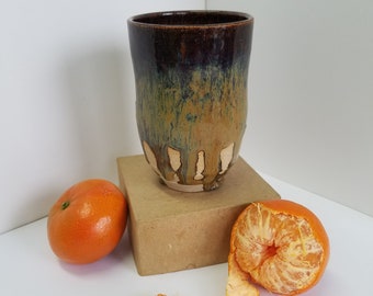 May include: A brown and green ceramic mug with a drip glaze sits on a brown cardboard box. Two mandarin oranges, one peeled, are on a white surface.