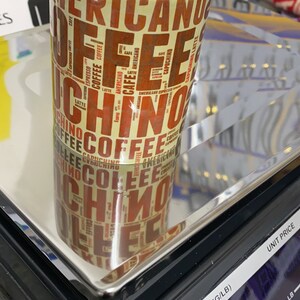 May include: A white ceramic mug with a brown and yellow design featuring the word "coffee" repeated in various fonts and sizes. The mug is sitting on a black digital scale with a silver platform.