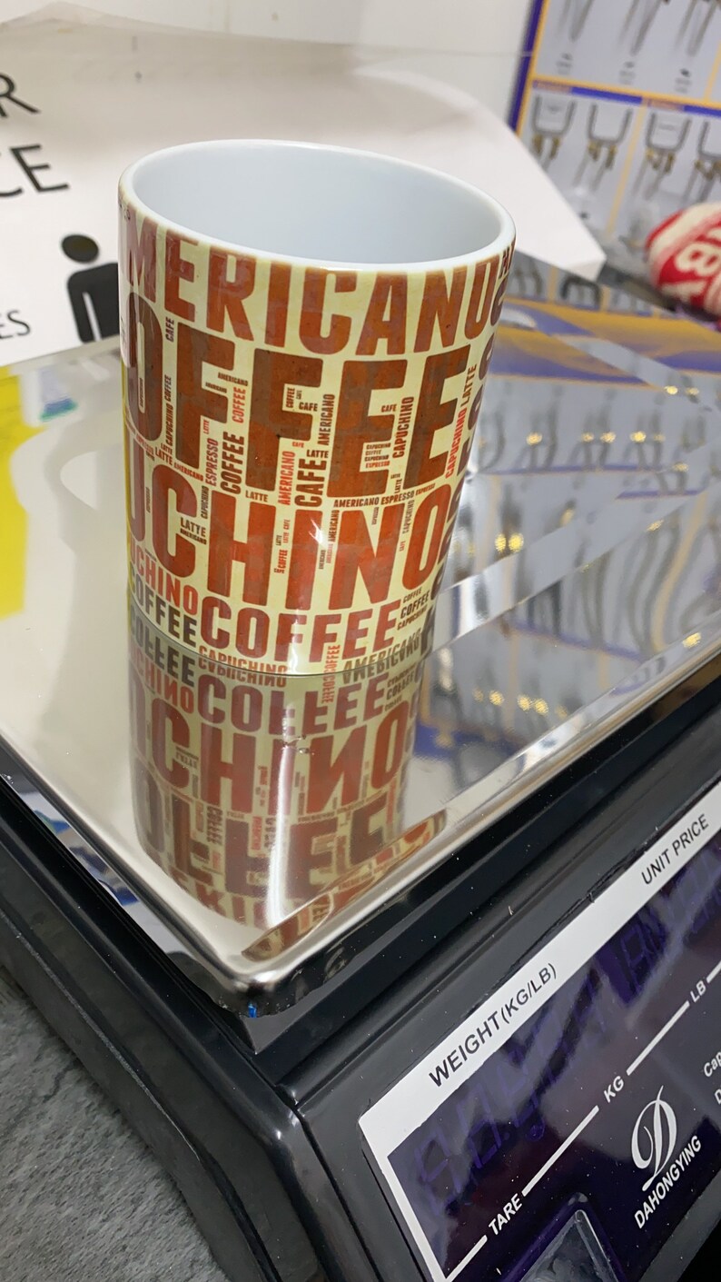 May include: A white ceramic mug with a brown and yellow design featuring the word "coffee" repeated in various fonts and sizes. The mug is sitting on a black digital scale with a silver platform.
